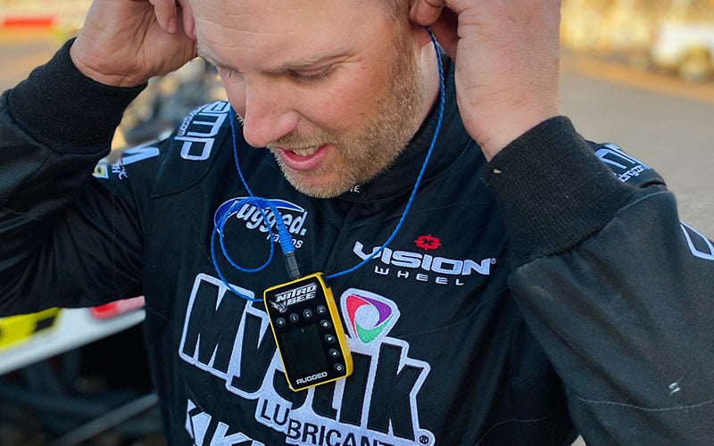 A close-up lifestyle shot of Cam Reimers wearing a black racing team jacket with 'Musk Lubricant' and 'Vision Wheel' logos, holding his ears as he wears a blue cord and a small yellow and black audio device around his neck.